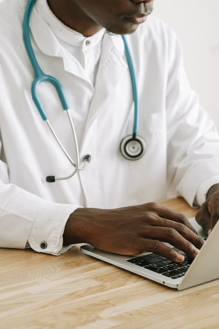 A professional doctor in a white coat with stethoscope typing on a laptop in an office setting.